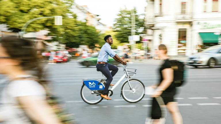 Image d'une scène de rue avec des piétons et un cycliste. Des arbres sont visibles à l'arrière-plan.